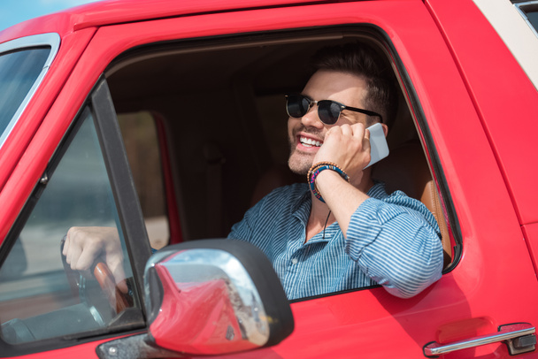 handsome cheerful man in sunglasses talking on smartphone in car during road trip - Фото, изображение