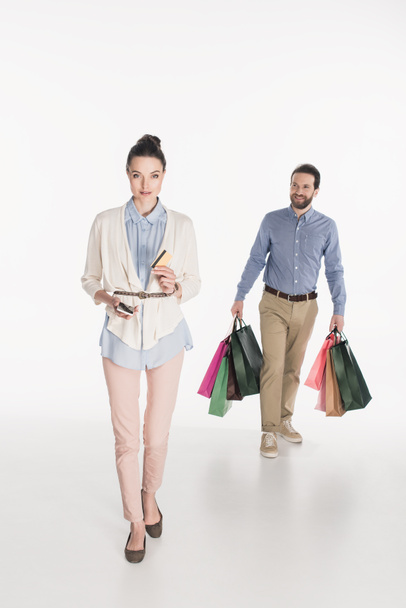 woman with credit card and smartphone looking at camera while husband carrying shopping bags isolated on white - Fotoğraf, Görsel