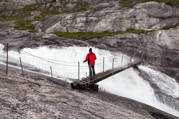 Viajante de pé sobre uma ponte suspensa sobre o rio Kinso no vale do Husedalen, Parque Nacional Hardangervidda, Noruega
 - Foto, Imagem