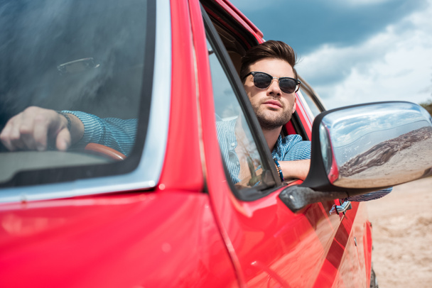 handsome man in sunglasses sitting in red car during road trip - Foto, afbeelding