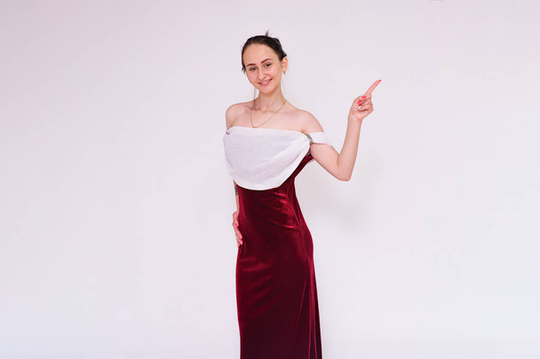 Look Here There is something interesting. Portrait of a beautiful brunette girl in a dress on a white background showing on the product. She is standing right in front of the camera smiling and looking happy - Photo, Image