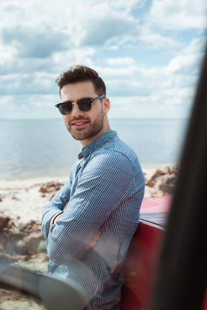 handsome smiling man standing at car near the sea during road trip - Fotó, kép
