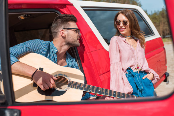 handsome man playing acoustic guitar near car with girlfriend - Photo, Image