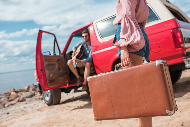 selective focus of girl with suitcase and boyfriend with guitar near red jeep on road trip - Photo, Image