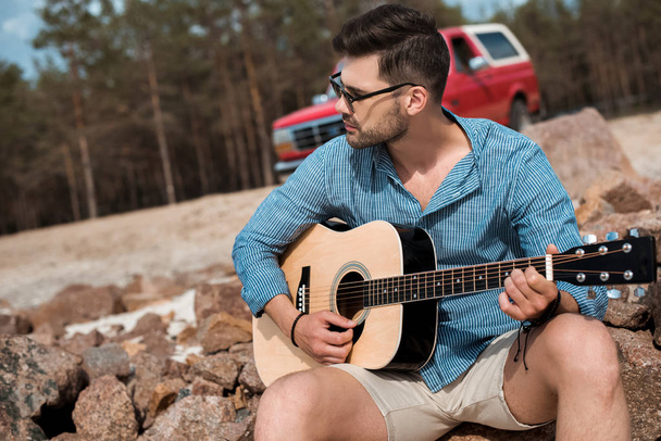 Junger Mann spielt Akustikgitarre im Freien, roter Jeep im Hintergrund - Foto, Bild