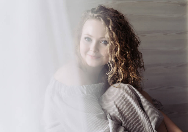 beautiful curly young woman sits by the window, enjoys relaxing and smiles - Fotografie, Obrázek