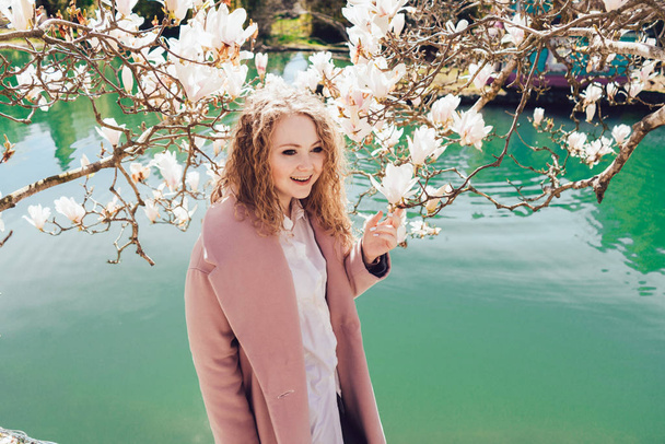 smiling young girl in pink coat posing in park next to pond, enjoying the sun and spring - Фото, изображение