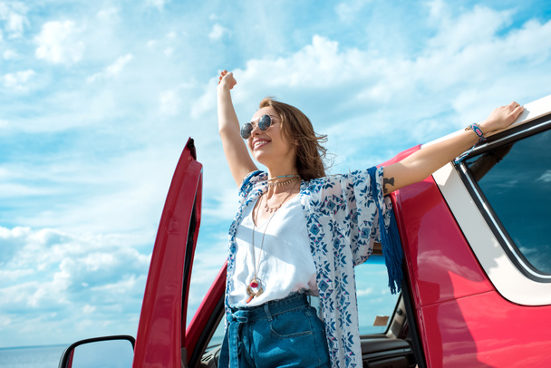 excited young woman in sunglasses standing near car on road trip - Foto, Bild