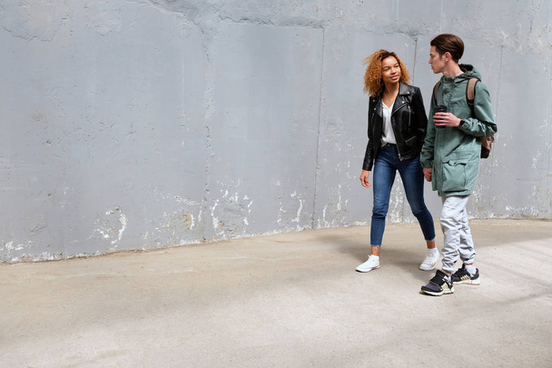 A black girl and a white guy look at each other, holding hands and walking along the street past a gray, shabby texture wall. Modern life style of international youth. Copy space for graffiti. - Foto, immagini