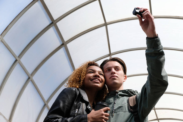 A modern szép afro-amerikai lány mosolyog, amikor együtt egy fehér fickó azt, hogy selfi a cselekvési kamera. Aranyos meg-diákok fényképezett össze belsejében egy tetőablak (kupola). - Fotó, kép