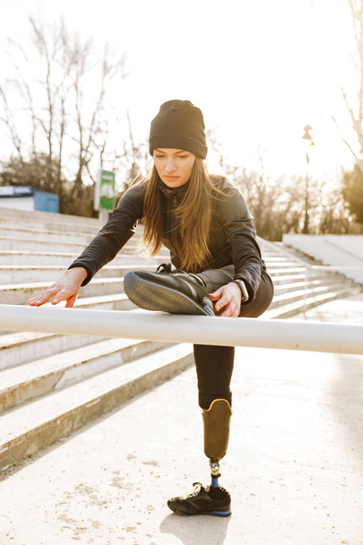 Image of disabled running girl in sportswear training and stretching prosthetic leg on railing in stadium outside - Φωτογραφία, εικόνα