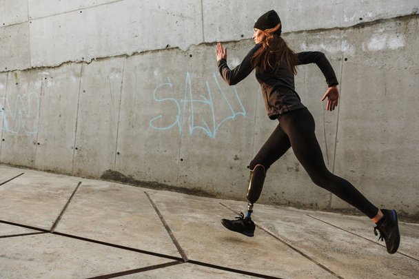Portrait of disabled athlete woman with prosthetic leg in tracksuit running outdoor along concrete wall - Фото, изображение