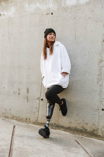 Photo of caucasian disabled girl with bionic leg in casual wear standing and smiling outdoor over concrete wall - Foto, Imagem