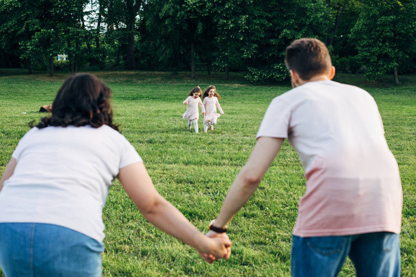 vista trasera de una pareja esperando a sus hijos gemelos de pie en la contemplación de un paisaje mientras sus dos hijos corriendo en la hierba
 - Foto, Imagen
