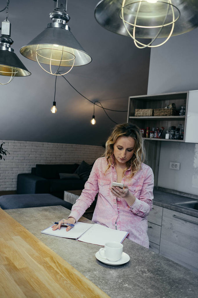 Beautiful young woman freelancer works in an apartment with phone and notebook. Business concep - 写真・画像