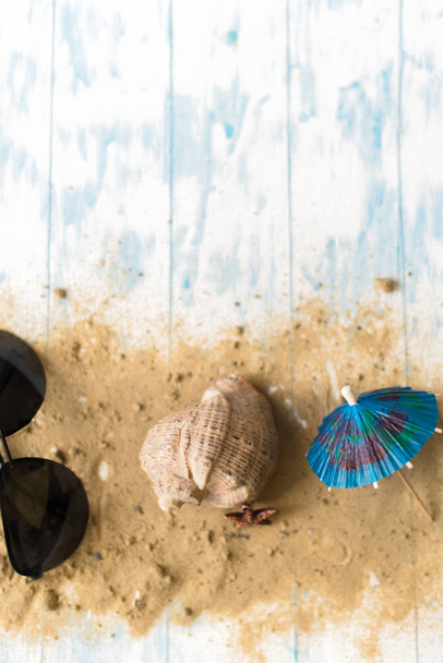 Beach still life. Black glasses with cocktail umbrellas, seashell on sand on a wooden background - Fotografie, Obrázek