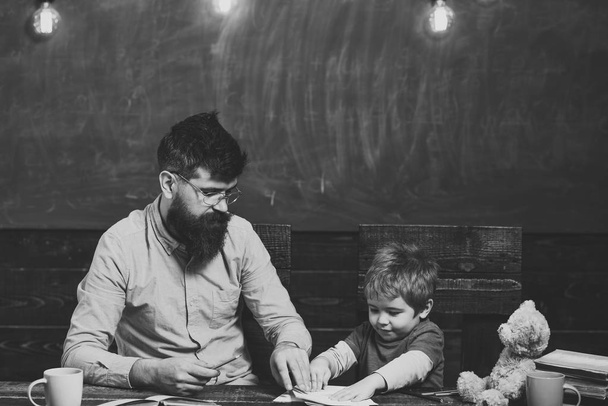 Happy kid having fun. Teacher and little kid sitting at desk with books and teddy bear. Schoolboy playing with copybook. Strict teacher in glasses in front of green board - Foto, Imagem