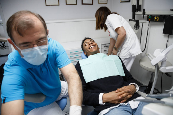 Lachender afrikanisch-amerikanischer Mann sitzt auf dem Stuhl des Zahnarztes in der Klinik und bereitet sich auf den Eingriff mit der Krankenschwester vor. - Foto, Bild