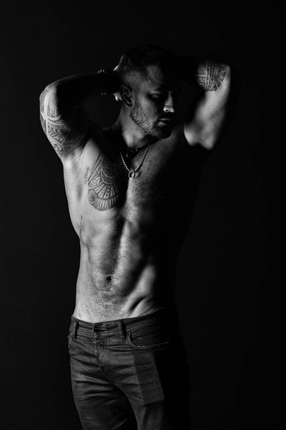Bodybuilder with biceps and triceps. Tattooed man show sexy muscular torso. Sportsman with six pack and ab. Fashion model with tattoo in jeans. Bodycare with fitness and sport, black and white - Foto, immagini