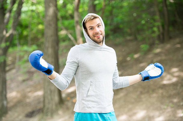 Sportsman boxer training with boxing gloves. Boxing concept. Man athlete on happy face with sport gloves, going to practice boxing, nature background. Boxer with hood on head ready for training - 写真・画像