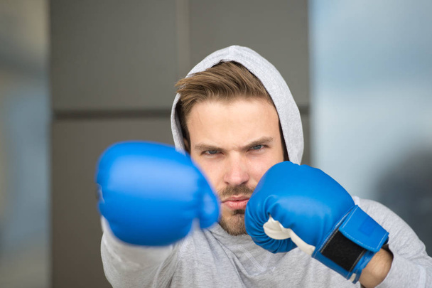 Boxing concept. Man athlete on concentrated face with sport gloves practicing boxing punch, urban background. Boxer with hood on head practices jab punch. Sportsman boxer training with boxing gloves - Фото, изображение