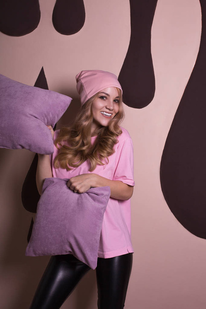 Blonde vrouw lachend met krullend haar spelen met kussens op een roze studio achtergrond - Foto, afbeelding