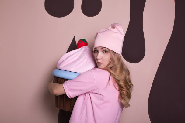 Hebzuchtige jonge blonde vrouw draagt roze t-shirt en cap, poserend met grote cupcake zoete decoratie - Foto, afbeelding