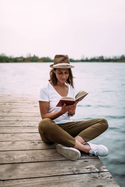 attractive young woman sitting near water on wooden jetty and reading book - Zdjęcie, obraz