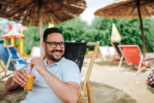 portrait of smiling young man drinking cocktail while sitting on beach chair, summer vacation concept  - Fotografie, Obrázek