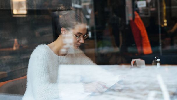 concentrated young girl freelancer in a white jacket and glasses is sitting in a cafe and writing a message on her smartphone - Fotoğraf, Görsel