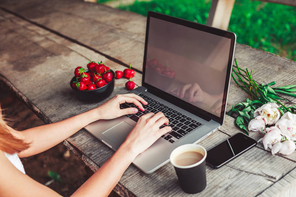 Young woman using and typing laptop computer at rough wooden table with coffee cup, strawberries, bouquet of peonies flowers, smartphone. Freelancer working in outdoor park. Toning - Foto, Bild