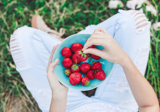 Meisje in spijkerbroek zit in zomer gras en een bord met aardbeien, de knieën en de handen zichtbaar te houden. Gezond ontbijt, schoon, eten, vegan food concept. Bovenaanzicht. Toning - Foto, afbeelding