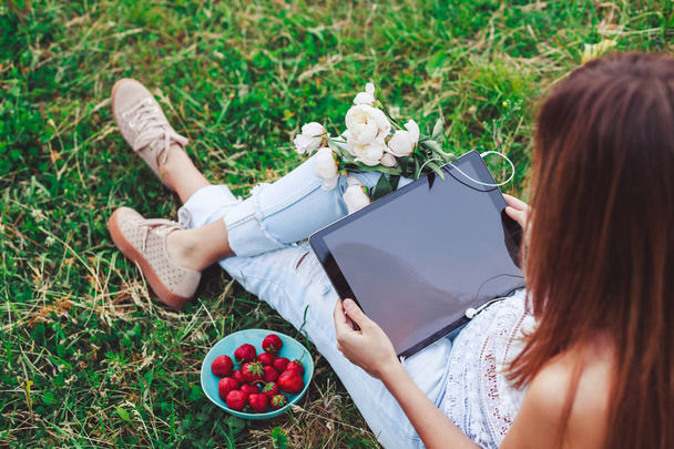 Mujer joven usando una tableta pc sentado en la hierba de verano con ramo de flores de peonías. Vacaciones de verano
 - Foto, imagen