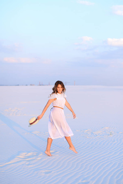 Young woman in white dress walking on sand and keeping hat. - Fotoğraf, Görsel