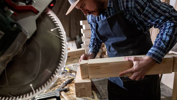 Ξυλουργός στο μικρό buiness ιδιοκτήτη εργάζεται σε εργαστήριο ξυλουργικής και ρούχα εργασίας, χρησιμοποιώντας ένα κυκλικό πριόνι για κόψιμο σε ένα ξύλινο, στο τραπέζι είναι ένα σφυρί και πολλά εργαλεία - Φωτογραφία, εικόνα