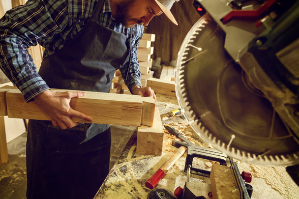 Плотник в рабочей одежде и владелец малого бизнеса работает в деревообрабатывающей мастерской, используя циркулярную пилу, чтобы прорезать дерево, на столе находится молоток и много инструментов
 - Фото, изображение