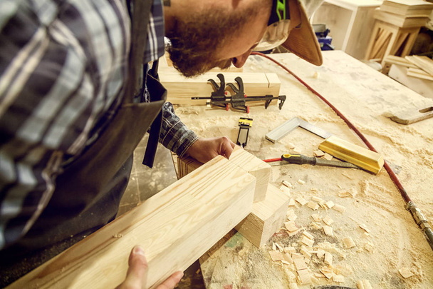 Ξυλουργός με ρούχα εργασίας κάνει ξυλοκατασκευών στην ξυλουργική. μικρό buiness ιδιοκτήτης λειτουργούν σε ξύλινη σανίδα στο εργαστήριο - Φωτογραφία, εικόνα