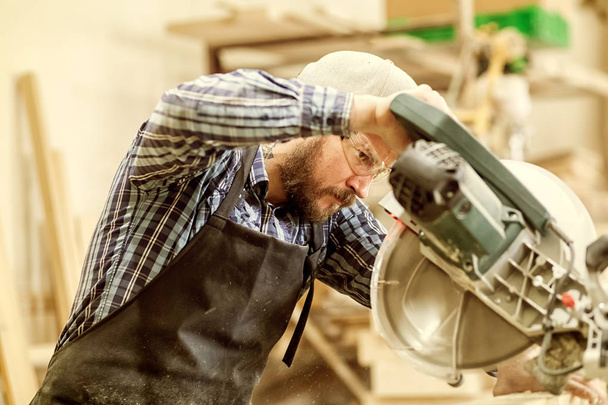 ein Tischler in Arbeitskleidung, der Holzarbeiten in Tischlerei verrichtet. Kleinunternehmer sägt in Werkstatt mit Kreissäge auf Holzbrett - Foto, Bild
