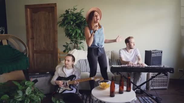 Pretty blonde vocalist from musical band is singing and dancing while her friends guitarist and keyboarder are playing guitar and keyboard and smiling. - Filmati, video
