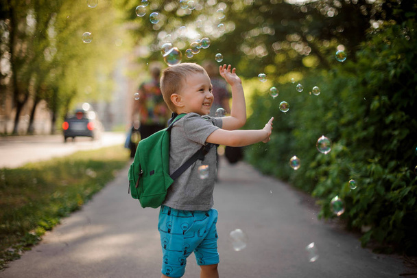 Happy little boy with a backpack playing with soap bubbles in green summer park on sunny day - Fotografie, Obrázek