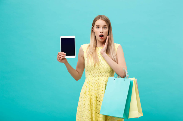 Aufgeregt schockiert gut aussehende junge Frau in gelbem Kleid, während sie Tablet und Einkaufstasche in der Hand hält. - Foto, Bild