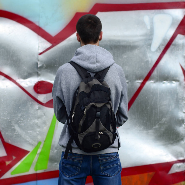 Un joven artista de graffiti con una bolsa negra mira a la pared con su graffiti en una pared. Concepto de arte urbano
. - Foto, Imagen
