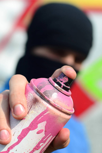 Ein junger Graffiti-Künstler in blauer Jacke und schwarzer Maske hält vor einem Hintergrund farbiger Graffiti-Zeichnungen eine Farbdose vor sich. Street Art und Vandalismus-Konzept. - Foto, Bild