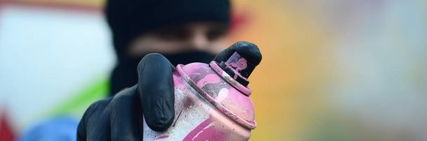 Ein junger Graffiti-Künstler in blauer Jacke und schwarzer Maske hält vor einem Hintergrund farbiger Graffiti-Zeichnungen eine Farbdose vor sich. Street Art und Vandalismus-Konzept. - Foto, Bild