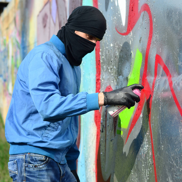 Um jovem hooligan com um rosto escondido pinta grafite em uma parede de metal. Conceito de vandalismo ilegal
. - Foto, Imagem