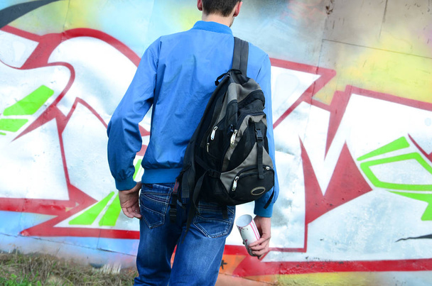 Un joven artista de graffiti con una bolsa negra mira a la pared con su graffiti en una pared. Concepto de arte urbano
. - Foto, Imagen