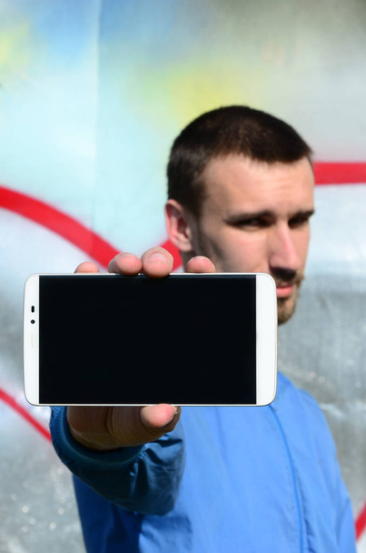 A graffiti művész azt bizonyítja, hogy a smartphone-val egy üres fekete képernyő, a háttérben a színes festett fal. Utcai művészeti koncepció. - Fotó, kép