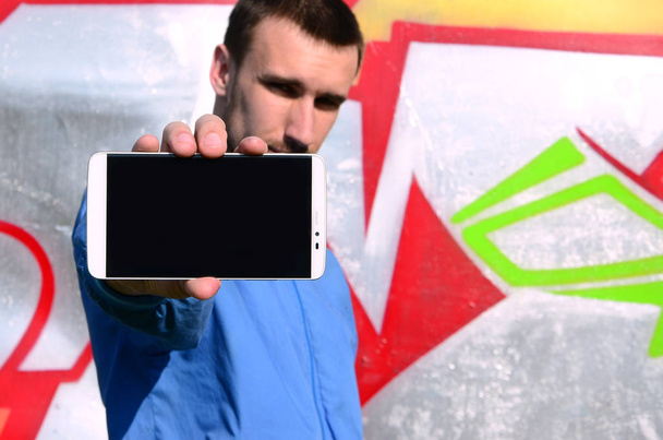El artista de graffiti muestra un smartphone con una pantalla negra vacía sobre el fondo de una colorida pared pintada. Concepto de arte urbano
. - Foto, Imagen