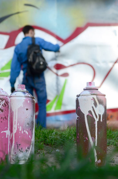 Unas cuantas latas de pintura usadas sobre el fondo del espacio con la pared sobre la que el joven dibuja un gran dibujo de graffiti. Arte moderno de dibujar paredes en graffiti
. - Foto, Imagen
