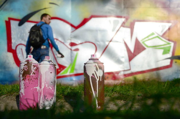 Kilka puszek farby używane na tle miejsca ze ścianą, na której młody facet rysuje duży graffiti rysunek. Nowoczesnej sztuki rysunku ściany graffiti. - Zdjęcie, obraz